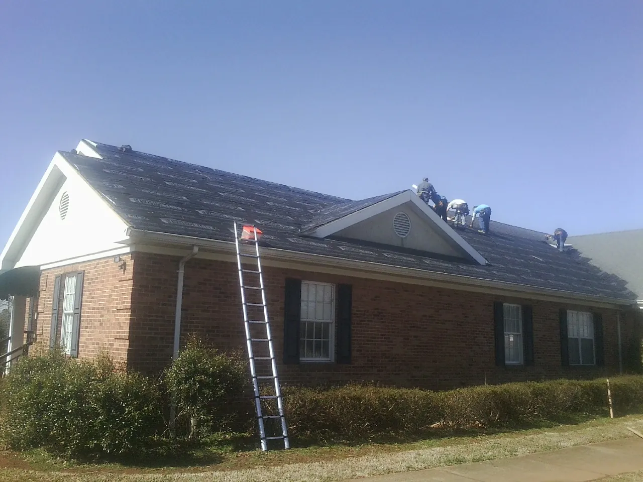 Crew tearing shingles off a brick ranch roof with a ladder set — Metal Roof Repair work by our crew in Constantine