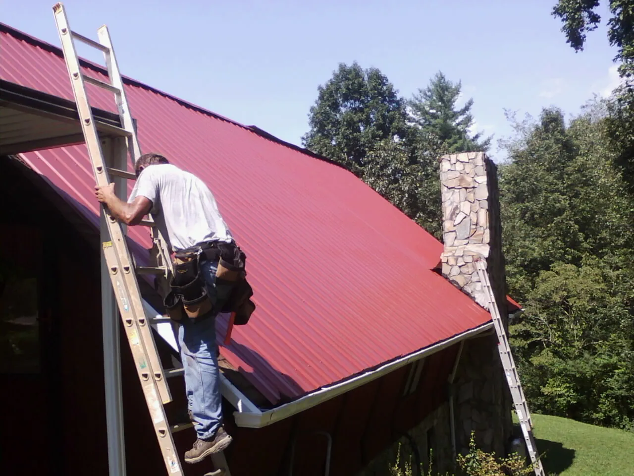 Roofer on a ladder beside a red metal roof and stone chimney — Soffit & Fascia Repair by Roofing Brothers in Constantine